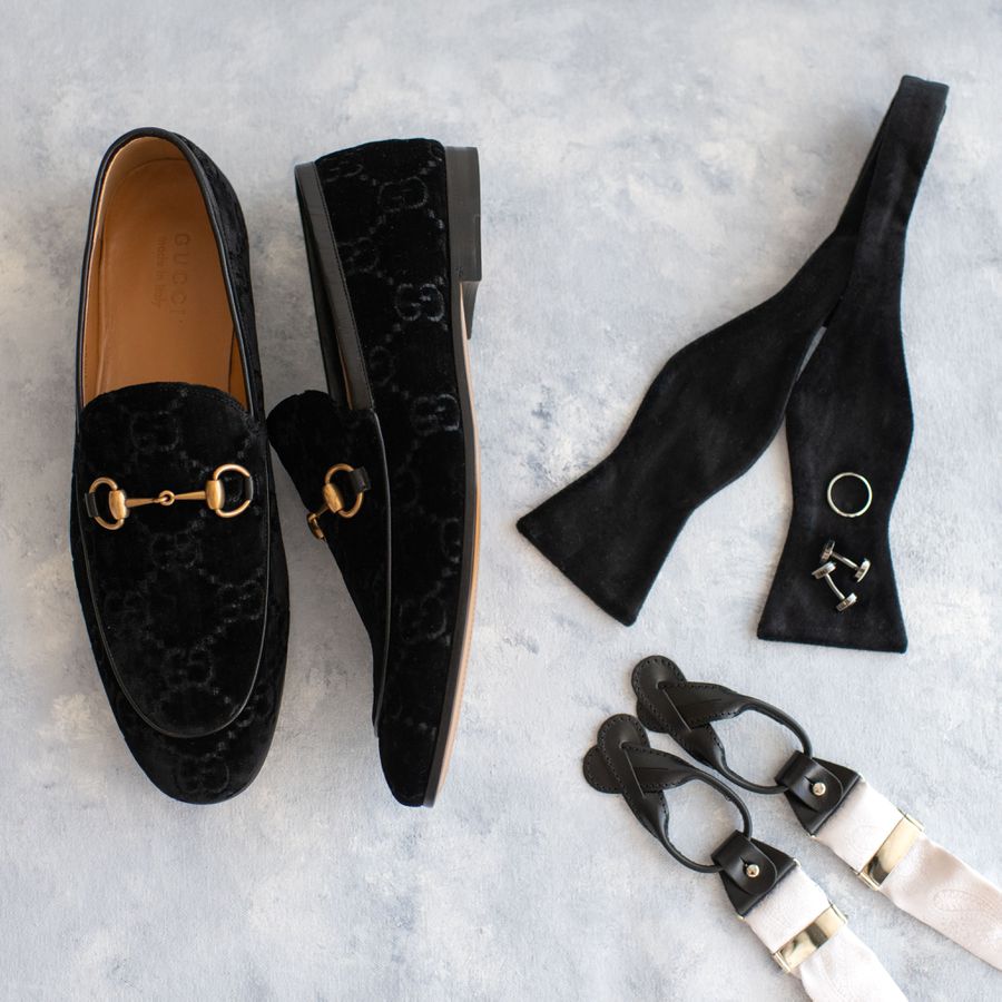 Velvet men's loafers displayed on a pale background next to an untied bowtie, cufflinks, and a wedding band