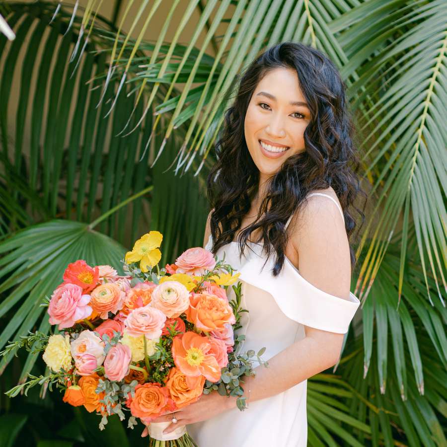 A bride with a lash lift and curly hair in an off-the-shoulder wedding dress holding a bouquet of peach, pink, and yellow ranunculus and roses
