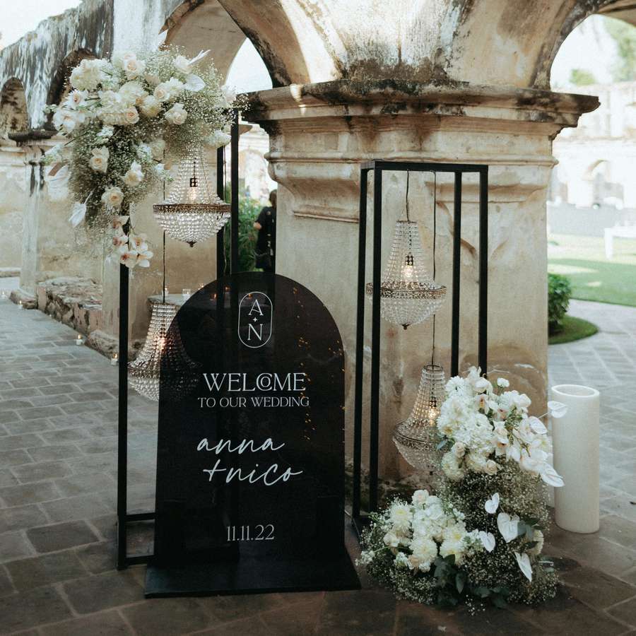 Black and white arched welcome sign decorated with crystal chandeliers and arrangements of roses and baby's breath