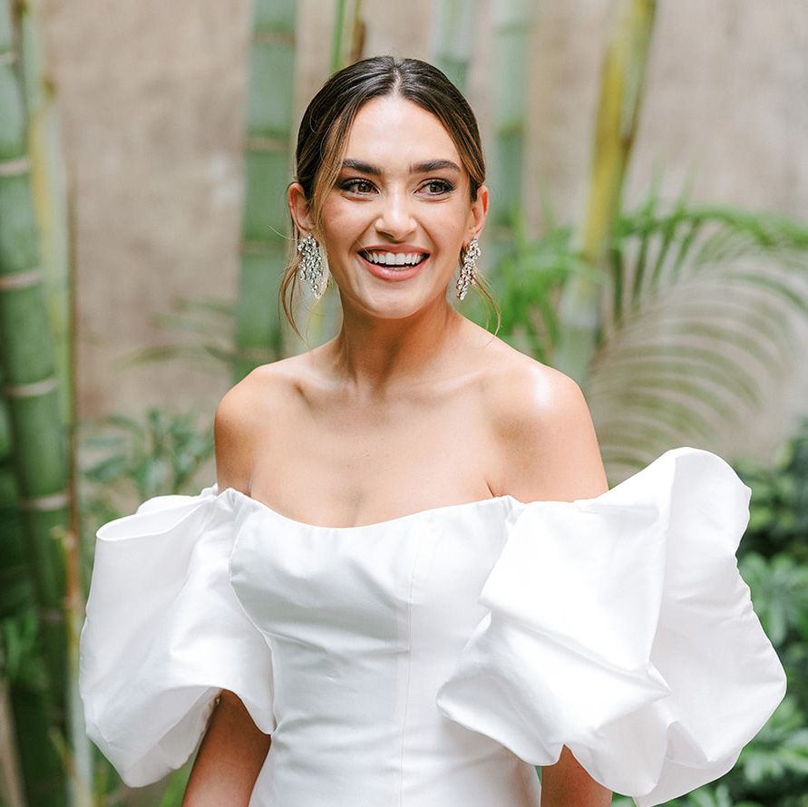 A bride smiling in front of bamboo wearing an off-the-shoulder wedding dress and chandelier earrings and showcasing defined eyebrows 